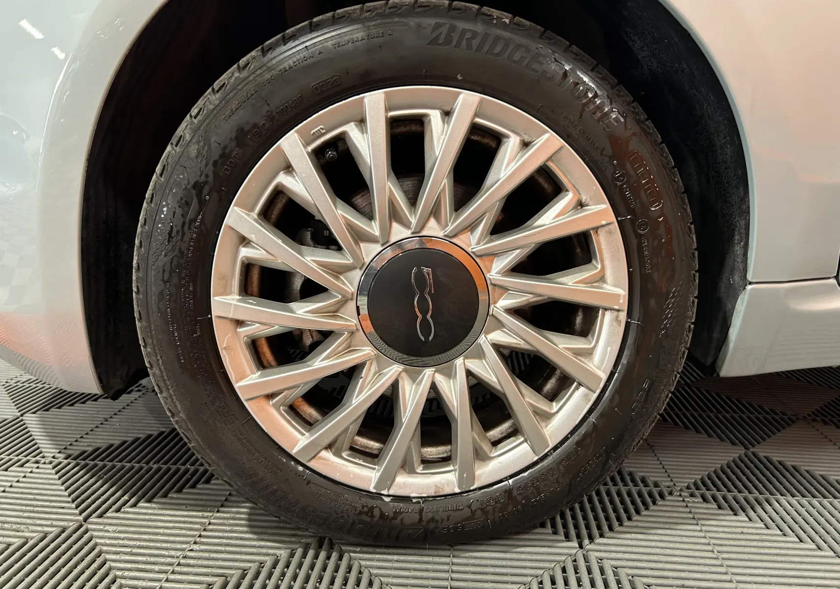 Gros plan sur la roue avant droite d'une FIAT 500 blanche avec jante argentée à motifs rayonnants.