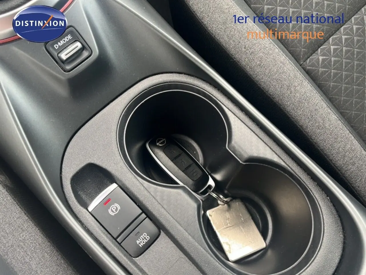 Gros plan sur la console centrale intérieure du Nissan Juke 2024, avec clé dans porte-gobelet et bouton frein à main électronique.