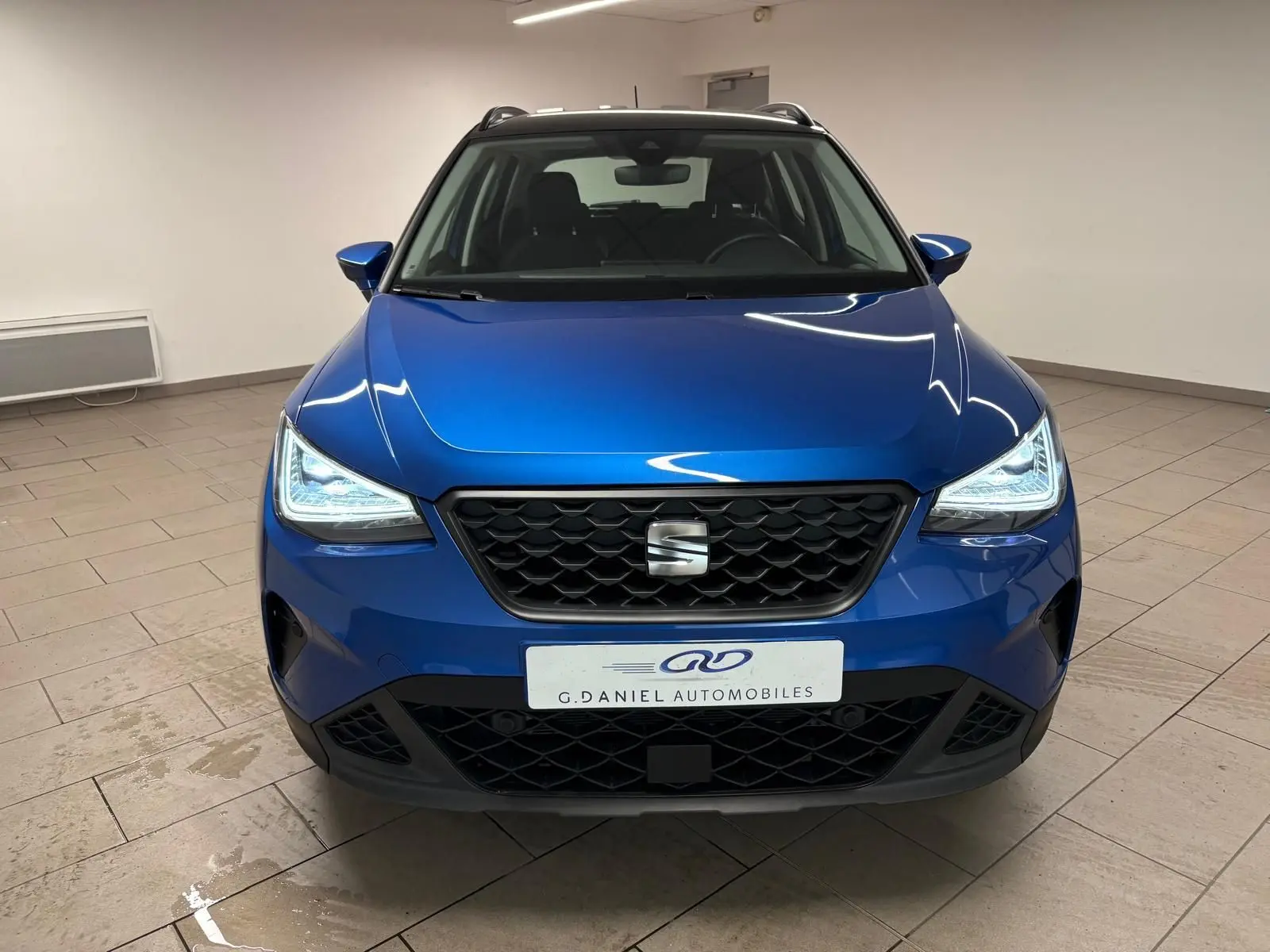 Vue avant d’un SEAT Arona bleu saphir avec toit noir, phares allumés et calandre noire en intérieur neutre.