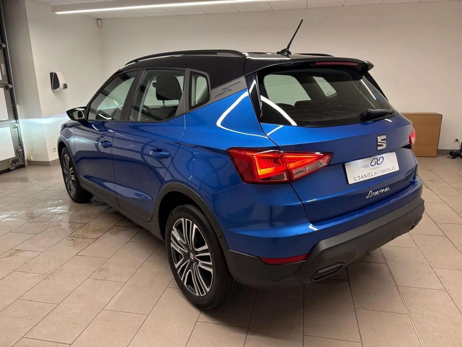 Vue 3/4 arrière droite d’un SEAT Arona 2025 bleu saphir avec toit noir, feux arrière allumés en intérieur.