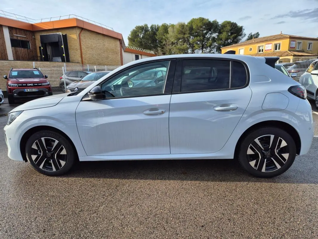 Profil droit d'une Peugeot 208 Hybrid 110 Allure blanche avec jantes alliage 16 pouces, vue en extérieur sur parking.