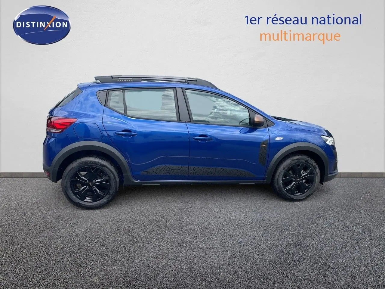 Profil latéral droit d'une Dacia Sandero Stepway 2024 bleu Iron Metal avec jantes noires et barres de toit.