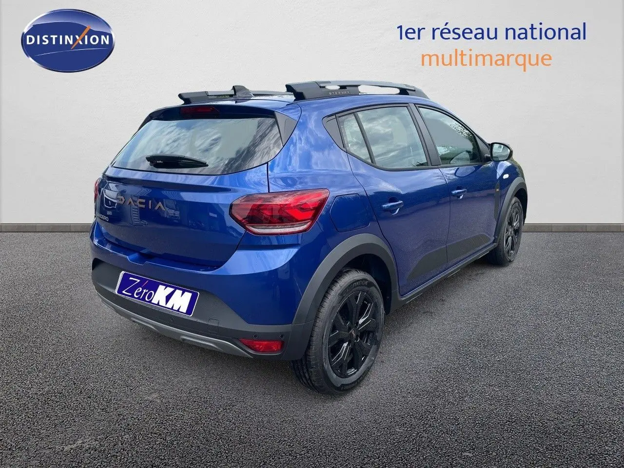 Vue 3/4 arrière droite d'une Dacia Sandero Stepway 2024 bleu iron metal avec jantes noires et barres de toit.