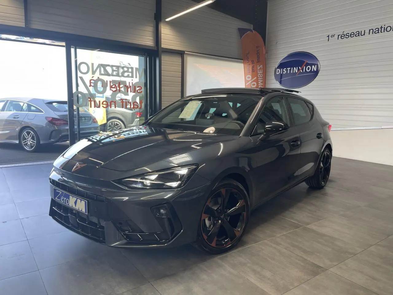 Vue 3/4 avant d'une CUPRA Leon gris magnétique avec jantes cuivrées et signature lumineuse Tri-LED en showroom.