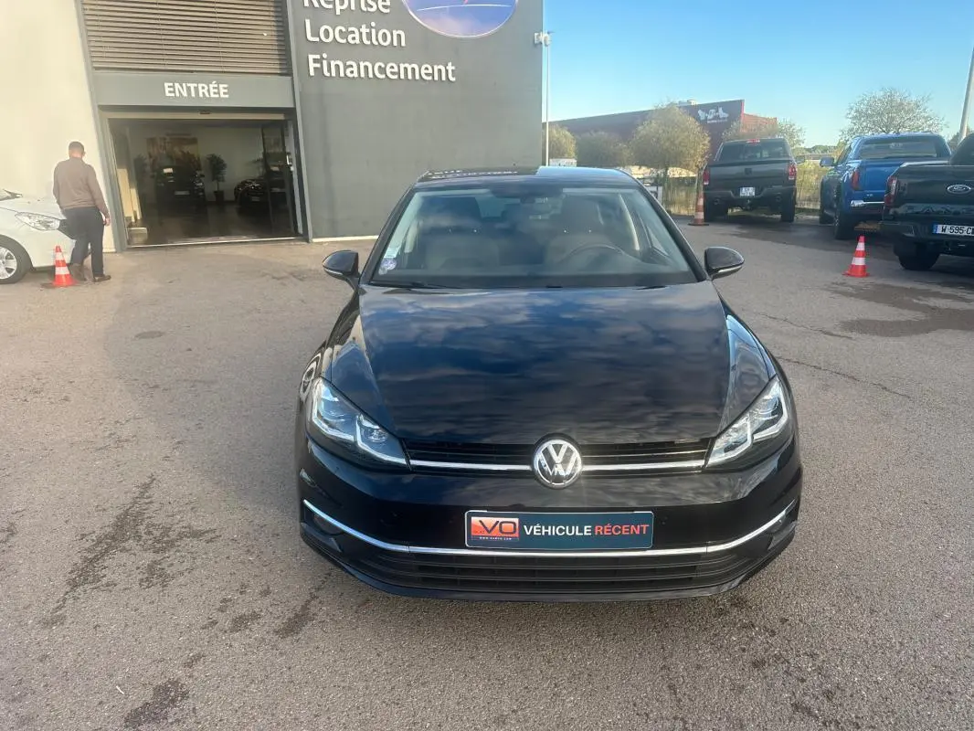 Vue de face d'une Volkswagen Golf noire 2018 avec phares allumés, stationnée devant un bâtiment commercial.