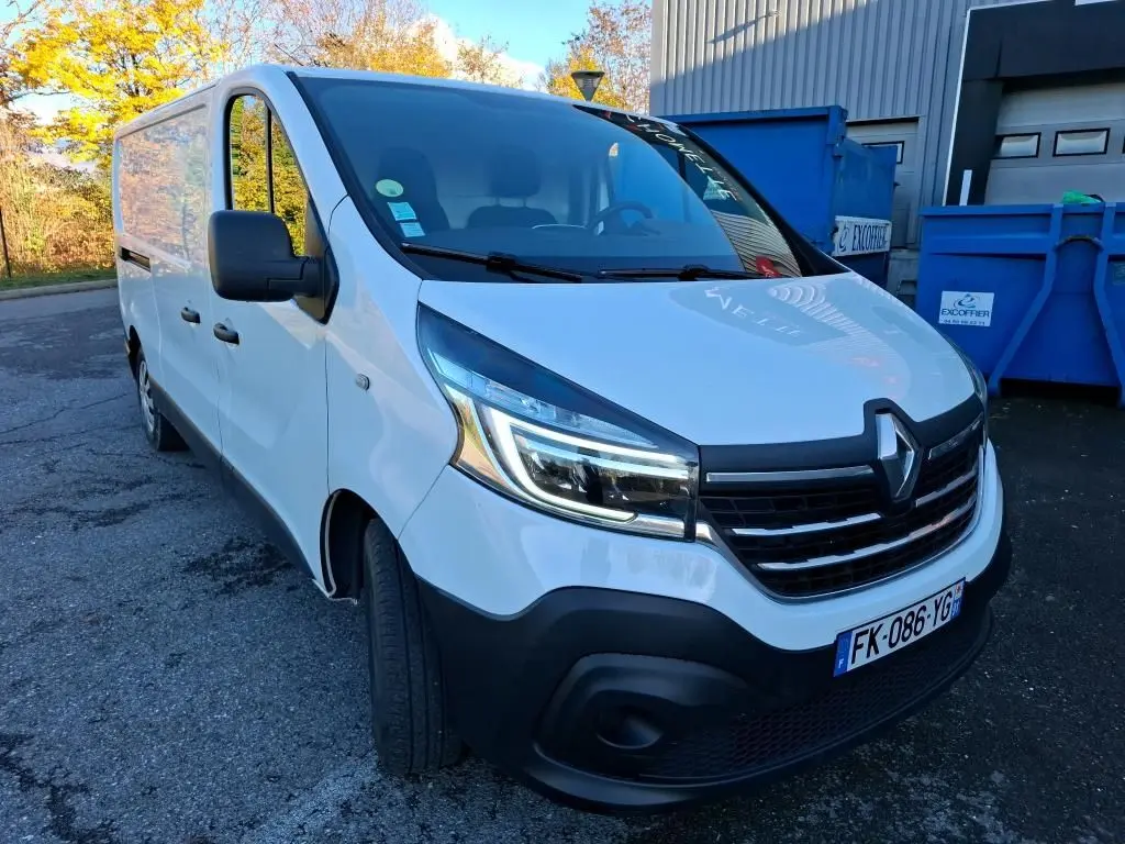 Vue 3/4 avant gauche d'un Renault Trafic Fourgon blanc de 2019 avec calandre noire et phares LED allumés.