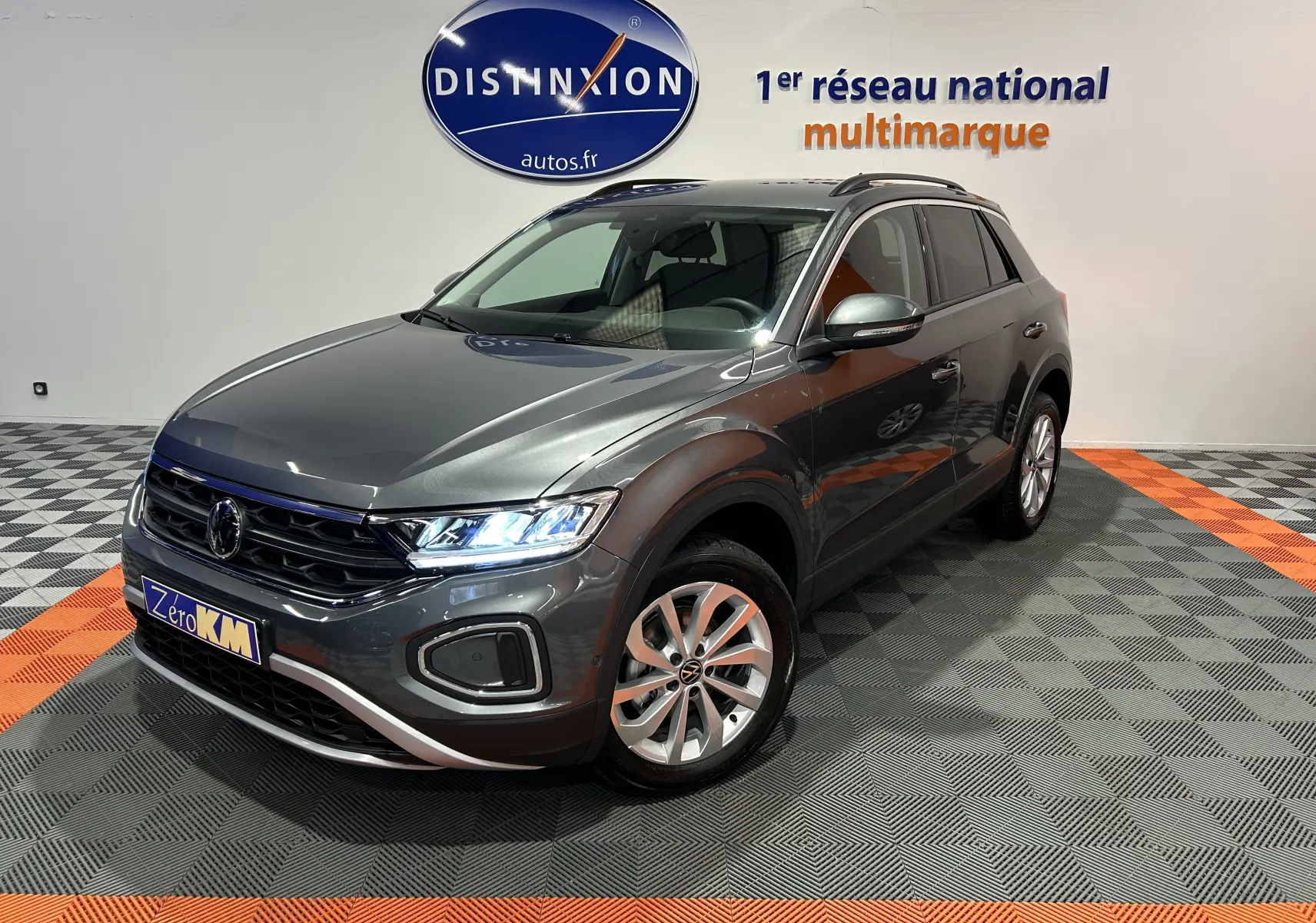 Volkswagen T-Roc gris Indium métal en 3/4 avant droit, avec feux allumés et jantes alliage visibles.