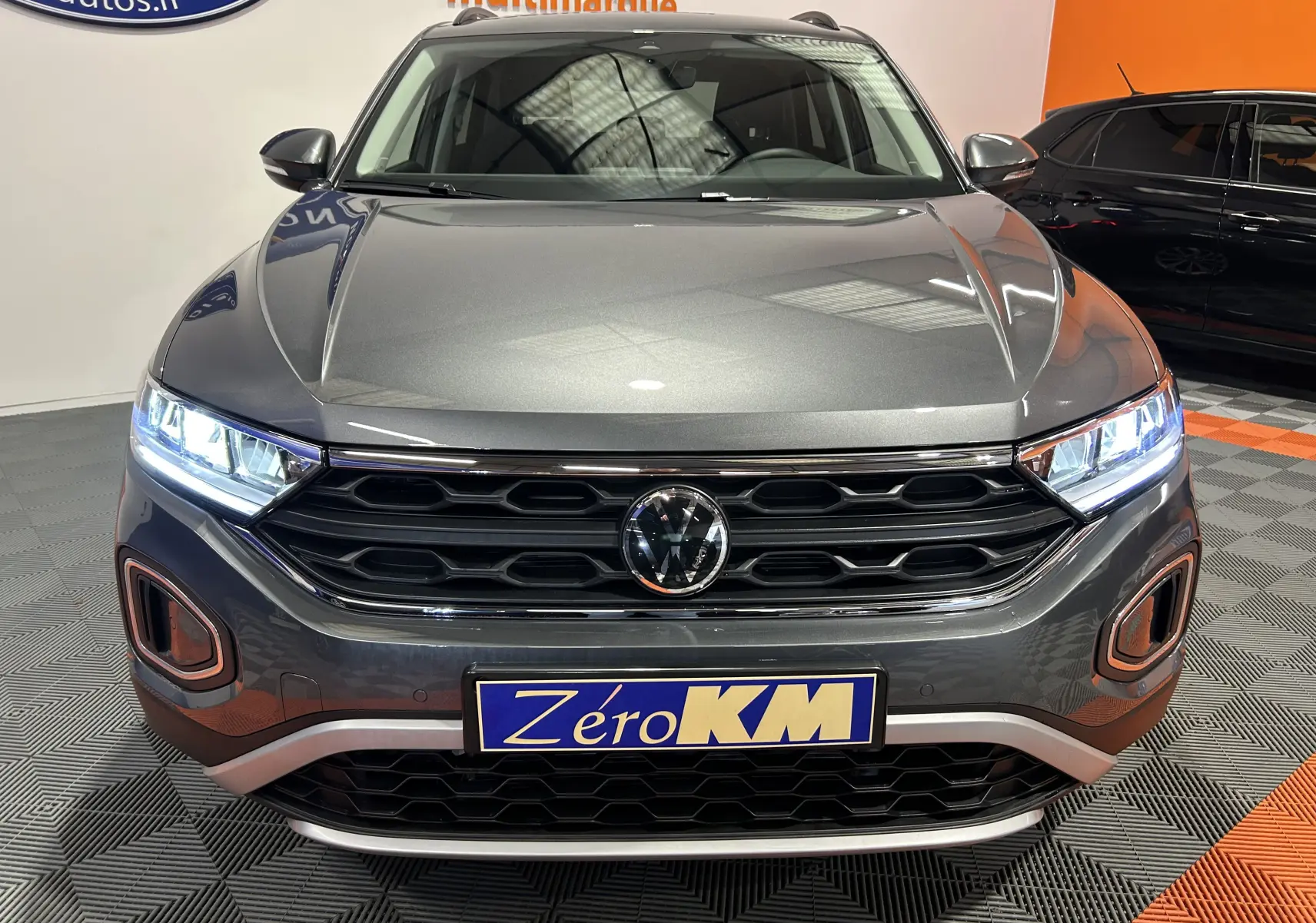 Vue frontale d’un Volkswagen T-Roc gris Indium Metal avec phares allumés et calandre noire en showroom.