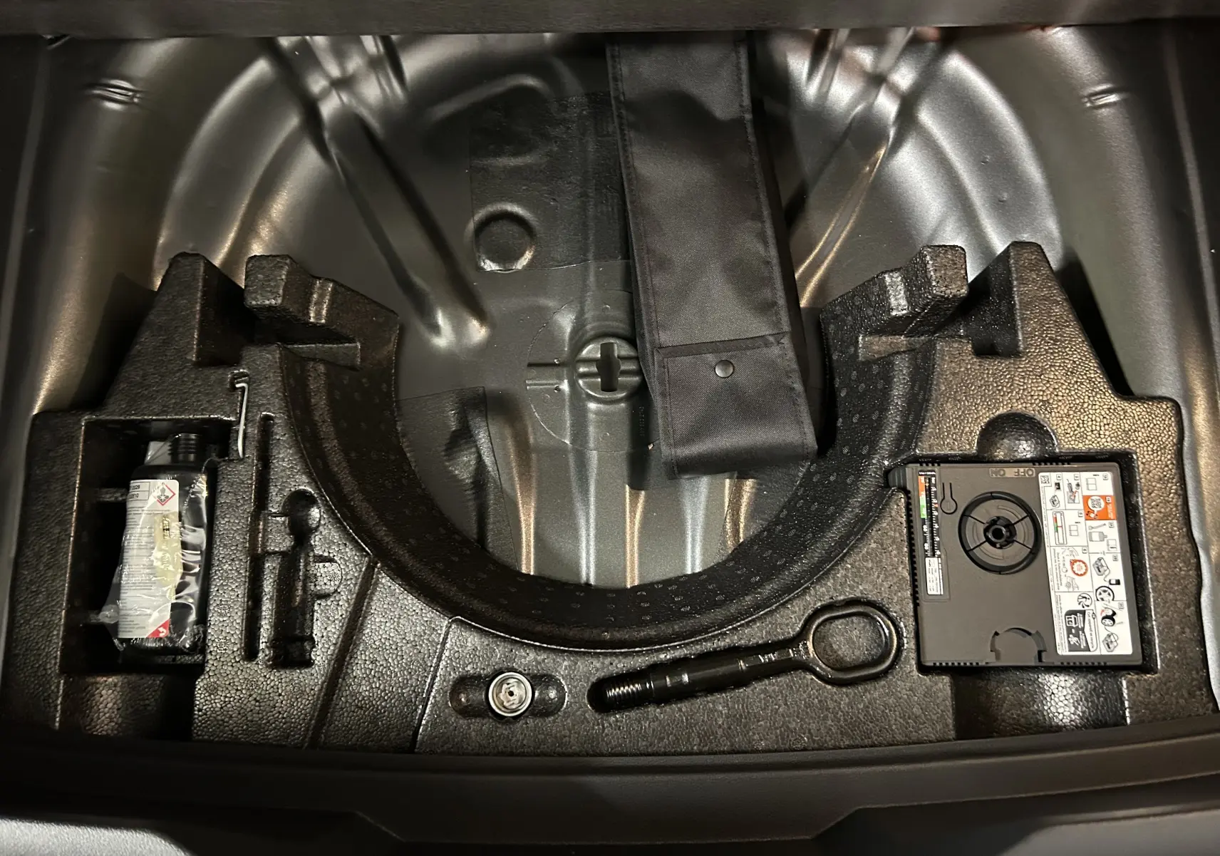 Vue plongeante sur le coffre ouvert du Volkswagen T-Roc gris Indium, montrant le rangement pour roue de secours et outils.