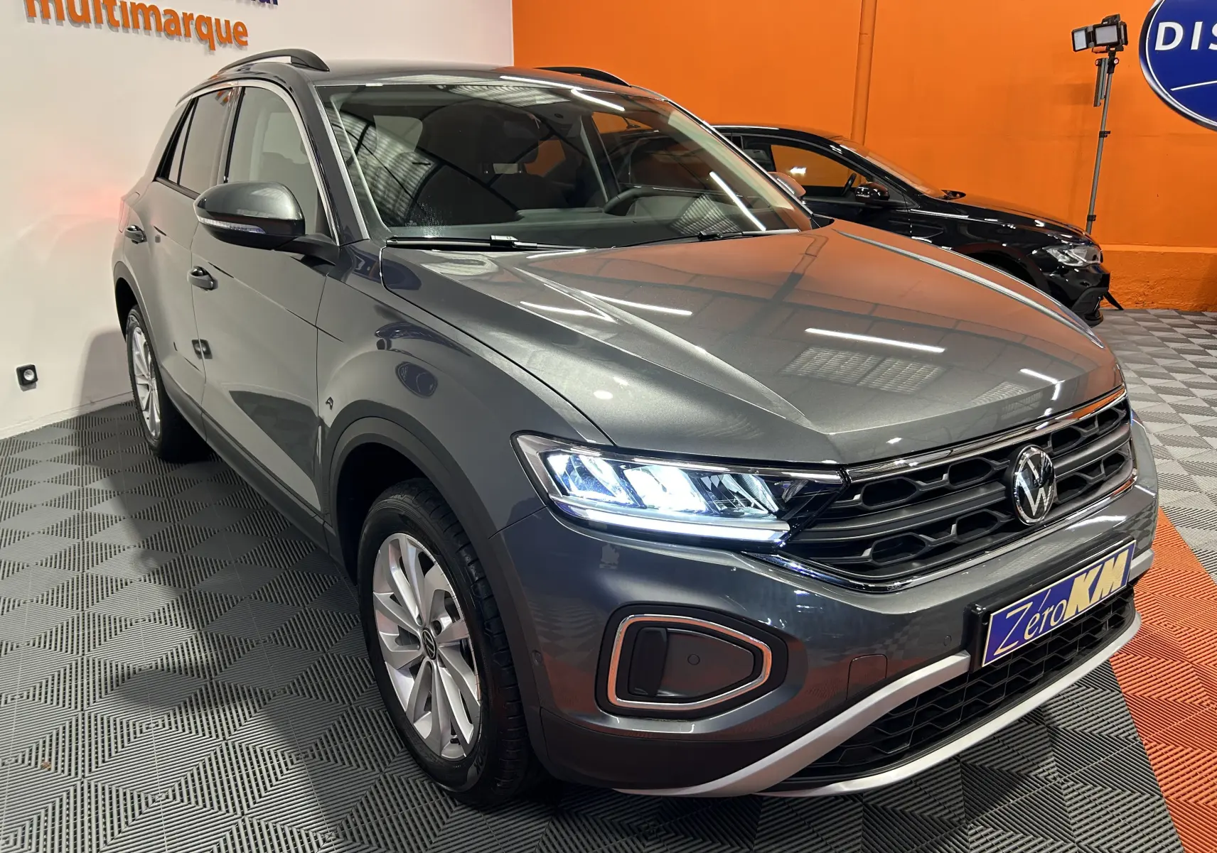 Volkswagen T-Roc gris Indium métal en 3/4 avant droit avec phares LED allumés dans un showroom.