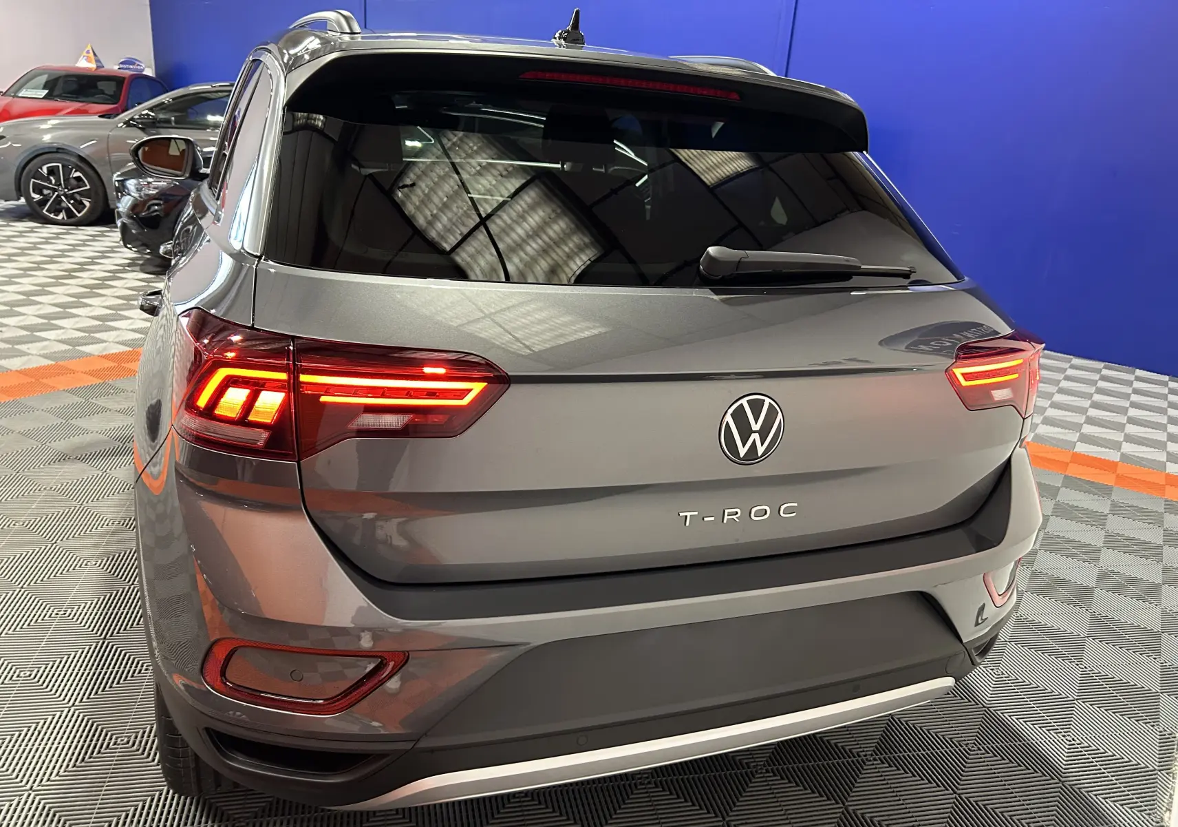 Vue 3/4 arrière d’un Volkswagen T-Roc gris Indium Metal avec feux arrière allumés dans un showroom.