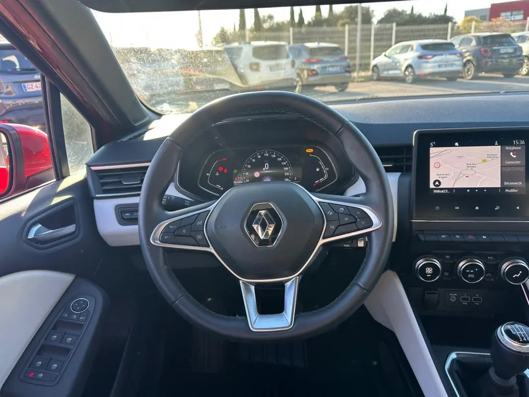Vue intérieure centrée sur le volant cuir et tableau de bord d'une Renault Clio rouge foncé, avec écran tactile et boîte manuelle.