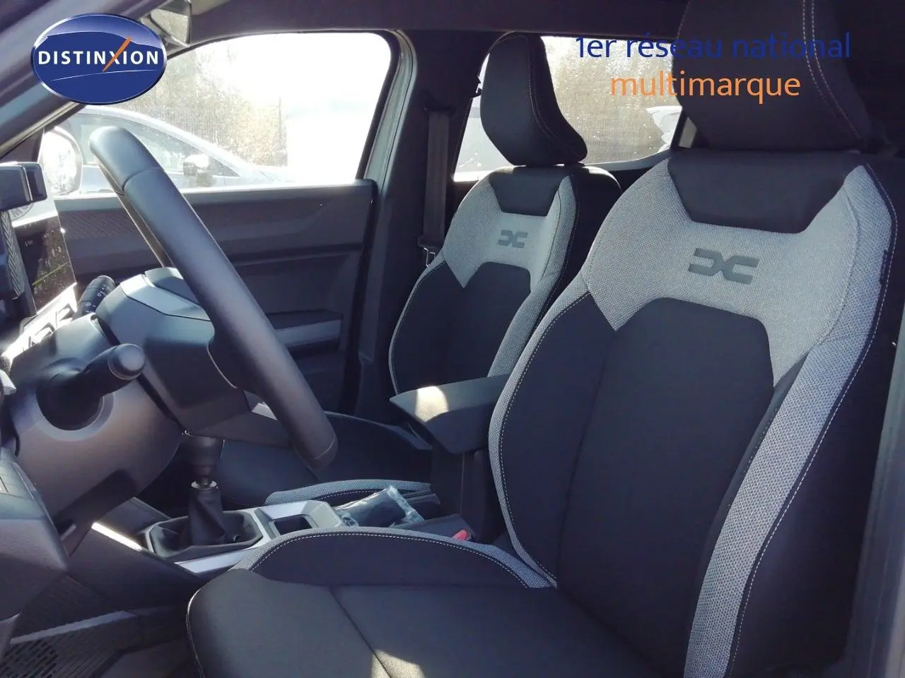 Vue intérieure côté conducteur du Dacia Duster 2026, sièges tissu gris et noir avec logo DC brodé, volant multifonction.