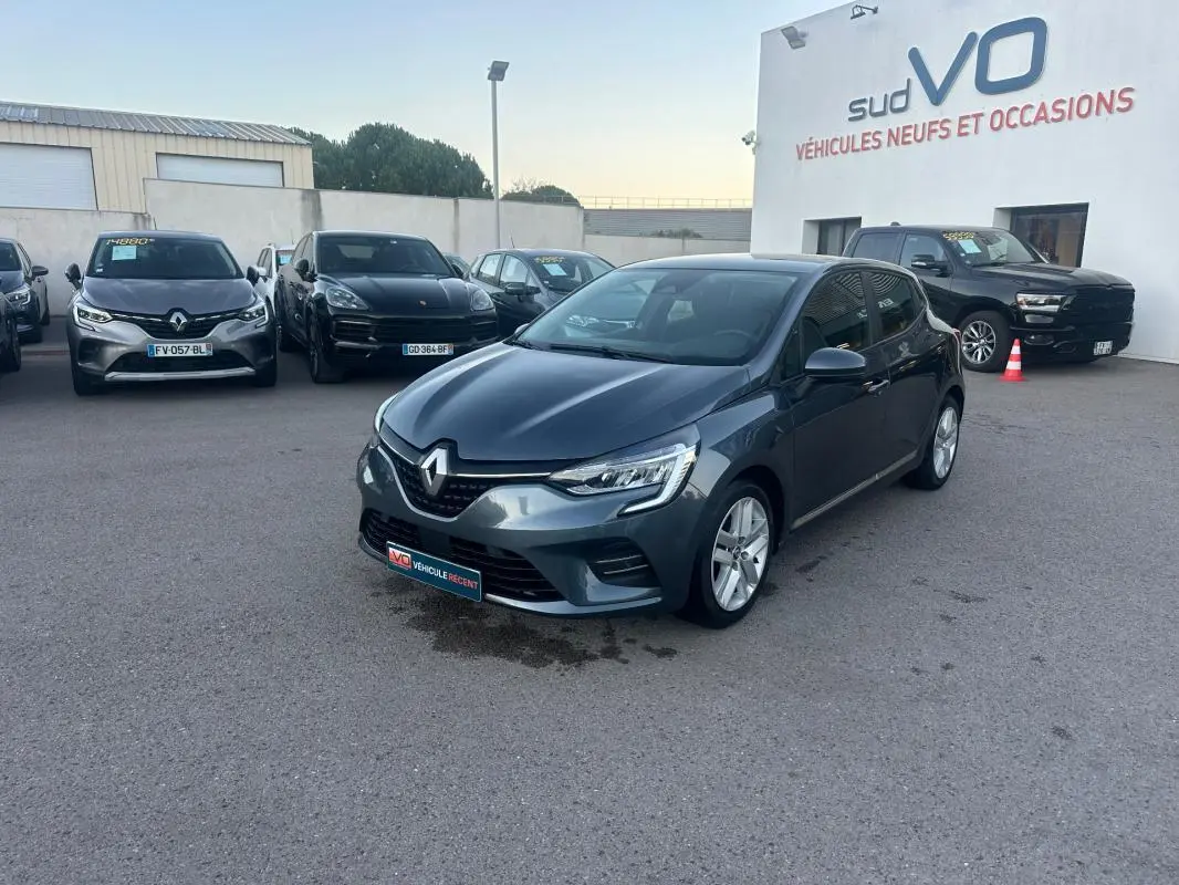 Renault Clio gris foncé vue 3/4 avant droit sur parking devant concession Sud VO, avec phares allumés et jantes alliage.