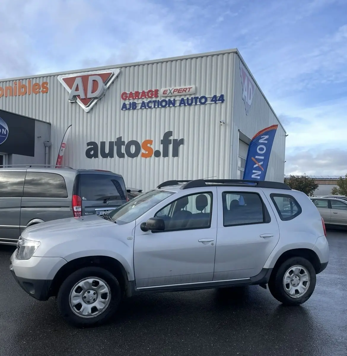 Vue de profil côté gauche d'un Dacia Duster gris avec barres de toit et enjoliveurs, stationné devant un garage.