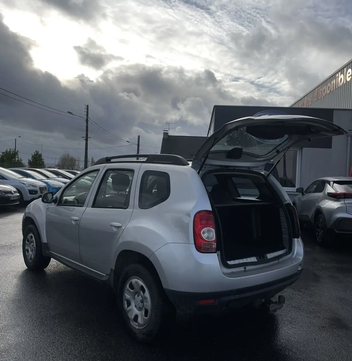 Vue 3/4 arrière droite d'un Dacia Duster gris 2011 avec hayon ouvert et attelage visible sur parking humide.