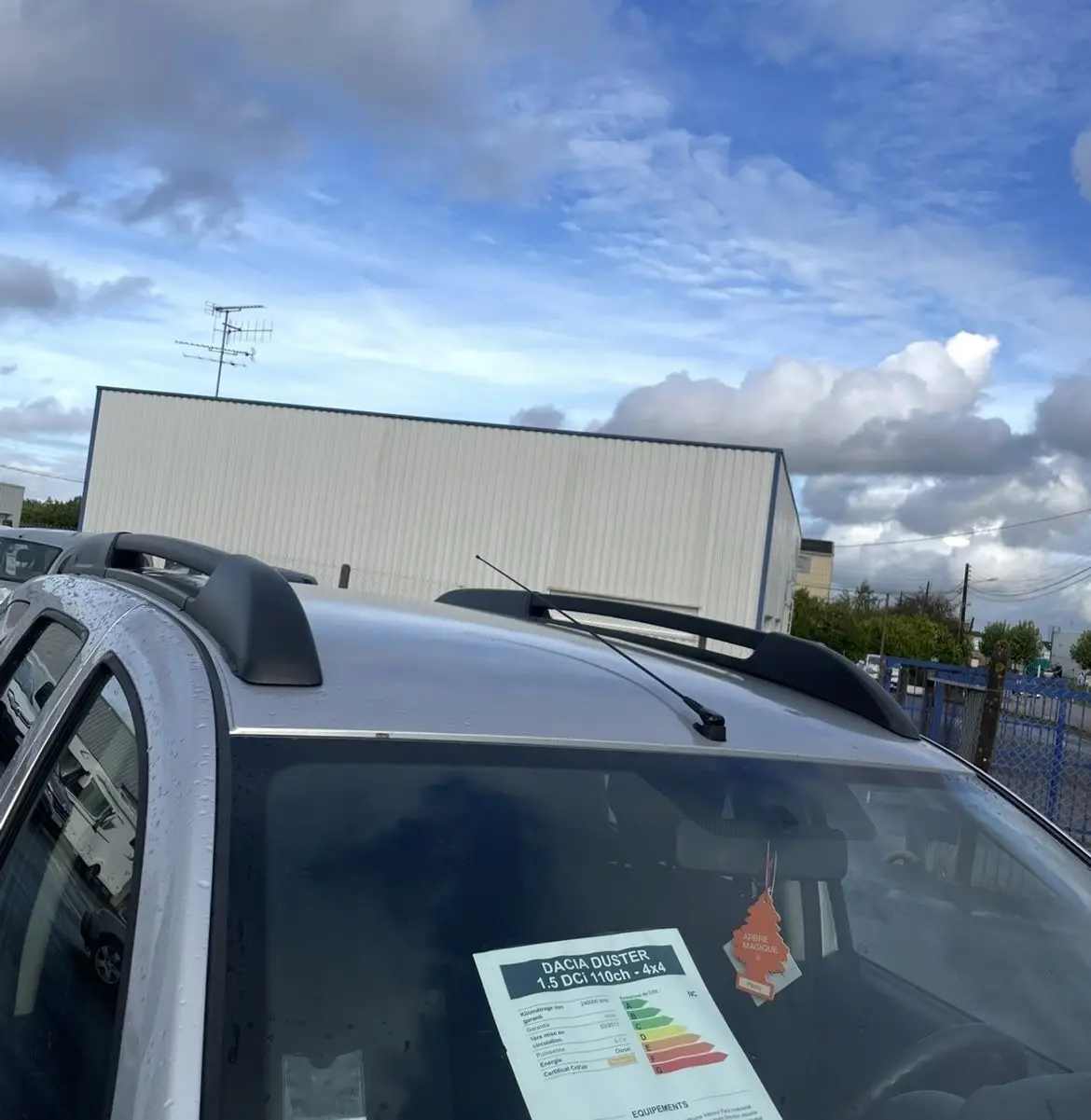 Vue 3/4 avant droit d'un Dacia Duster gris 2011 avec barres de toit et étiquette de vente visible sur le pare-brise.