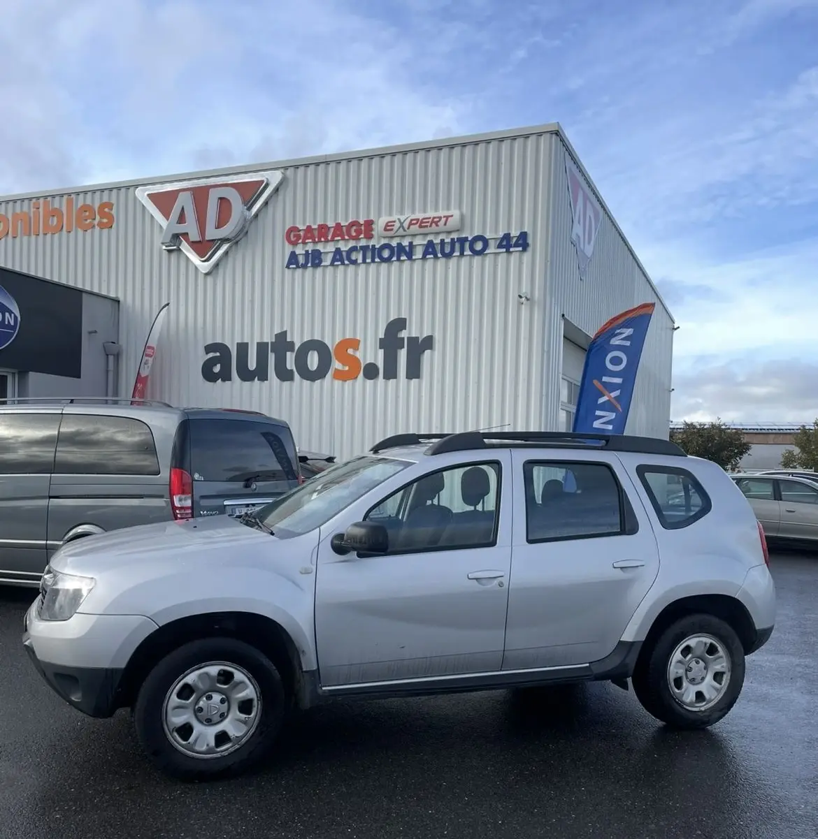 Profil côté gauche d'un Dacia Duster gris 2011 avec barres de toit et enjoliveurs, stationné devant un garage autos.fr.