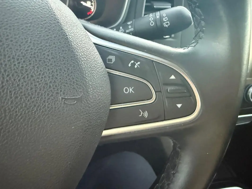 Gros plan sur les commandes du volant cuir noir de la Renault Mégane IV Berline gris clair, avec boutons multifonctions et chrome.