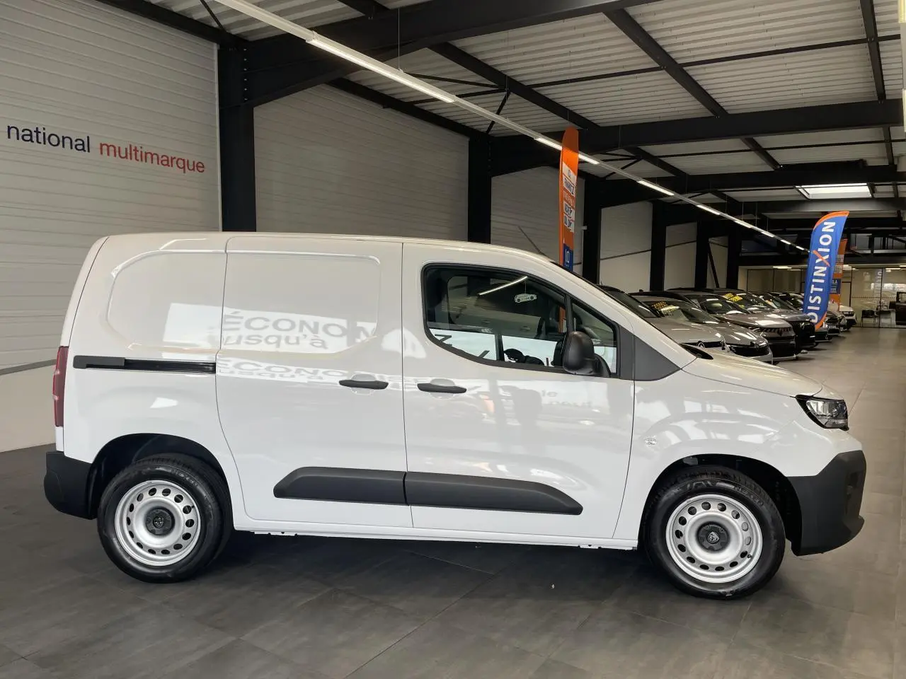 Vue de profil côté droit d’un Peugeot Partner blanc en fourgon dans un showroom, avec jantes tôles grises et porte latérale coulissante.
