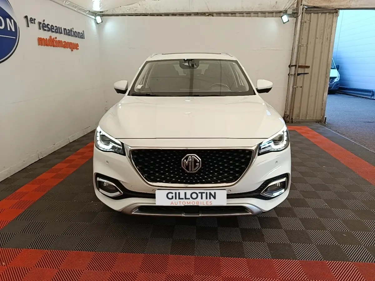 Vue frontale d’un MG Motor EHS blanc 2023 avec calandre noire et phares LED allumés dans un garage.