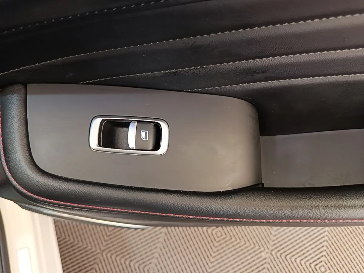 Gros plan sur le bouton de commande de la vitre électrique côté conducteur, entouré de cuir noir avec surpiqûres rouges.
