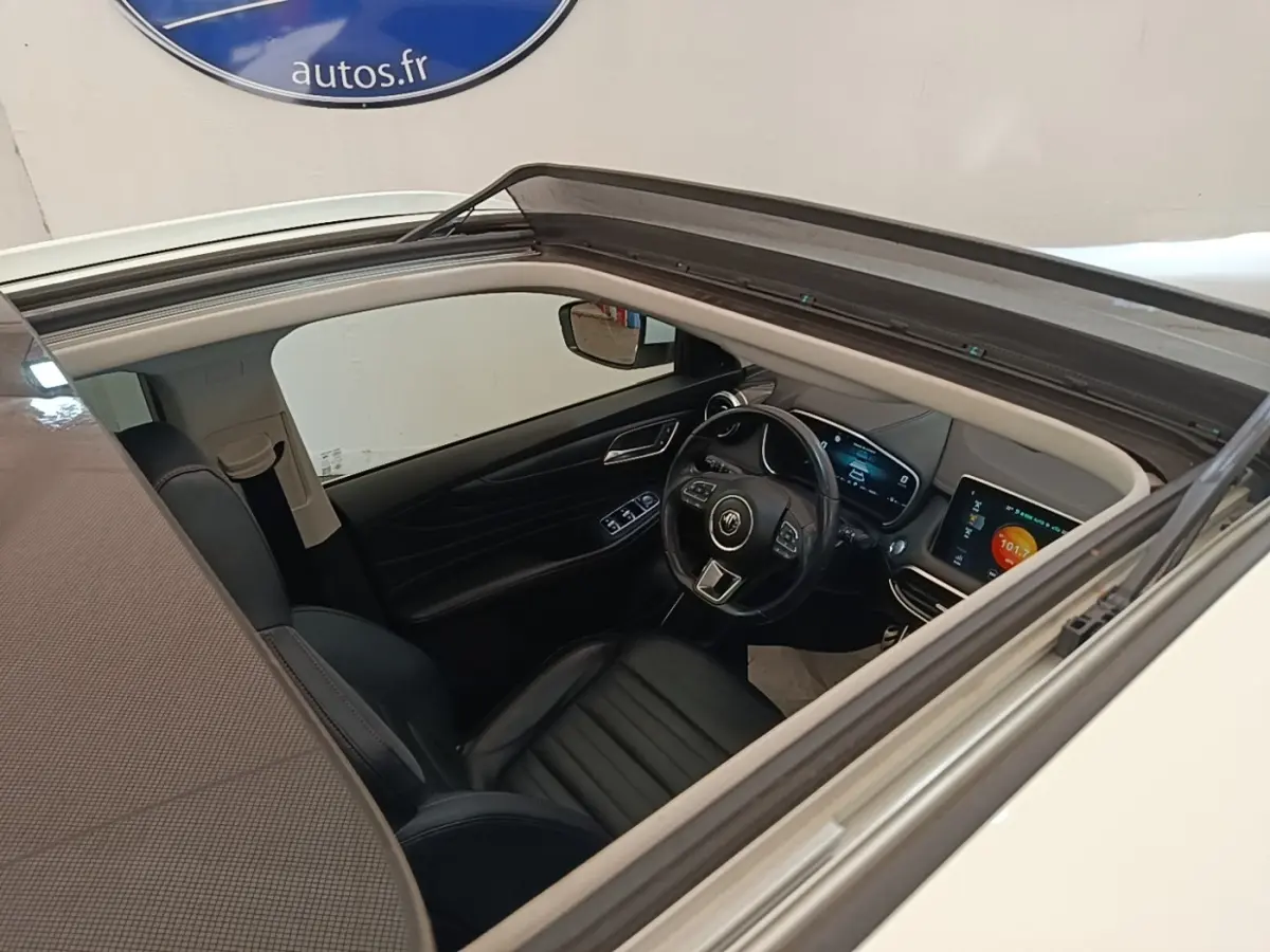Vue intérieure par le toit ouvrant du poste de conduite du MG Motor EHS blanc avec tableau de bord digital et volant cuir.
