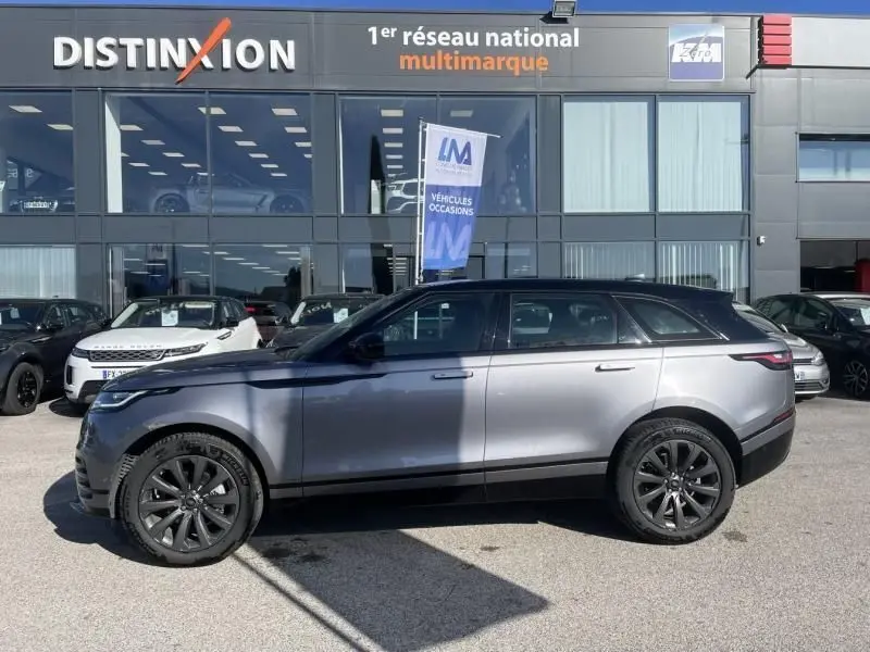 Vue de profil côté gauche d'un Range Rover Velar gris métal 2020 avec jantes noires, devant un concessionnaire multimarque.