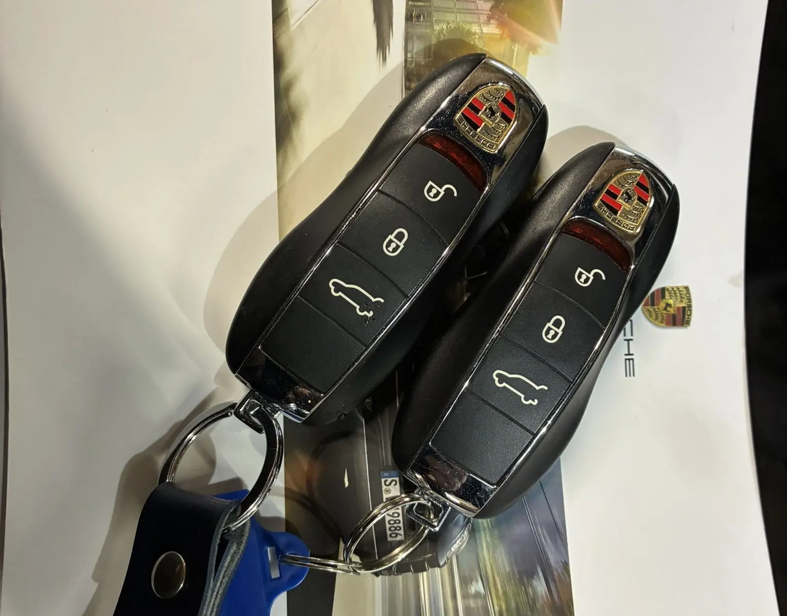 Gros plan sur deux clés noires Porsche Macan GTS posées sur une brochure avec logo et photo floue d'une voiture en mouvement.