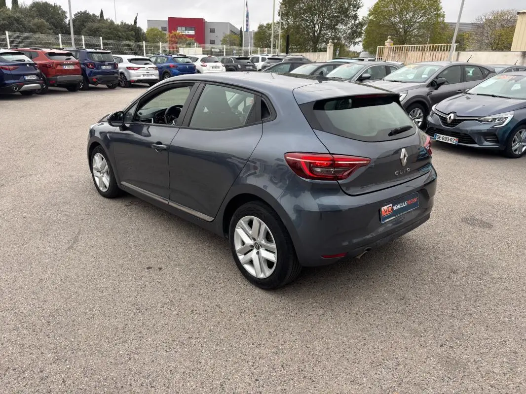 Renault Clio gris foncé vue 3/4 arrière côté gauche, avec feux arrière allumés sur parking extérieur.