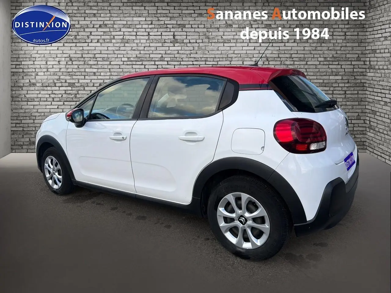 Vue 3/4 arrière droite de la Citroën C3 blanche avec toit rouge et jantes argentées, stationnée devant un mur en briques.
