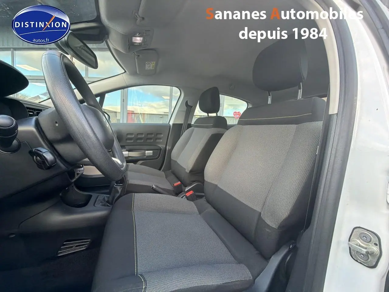 Vue intérieure côté conducteur de la Citroën C3 blanche 2018, sièges tissu gris avec surpiqûres jaunes visibles.