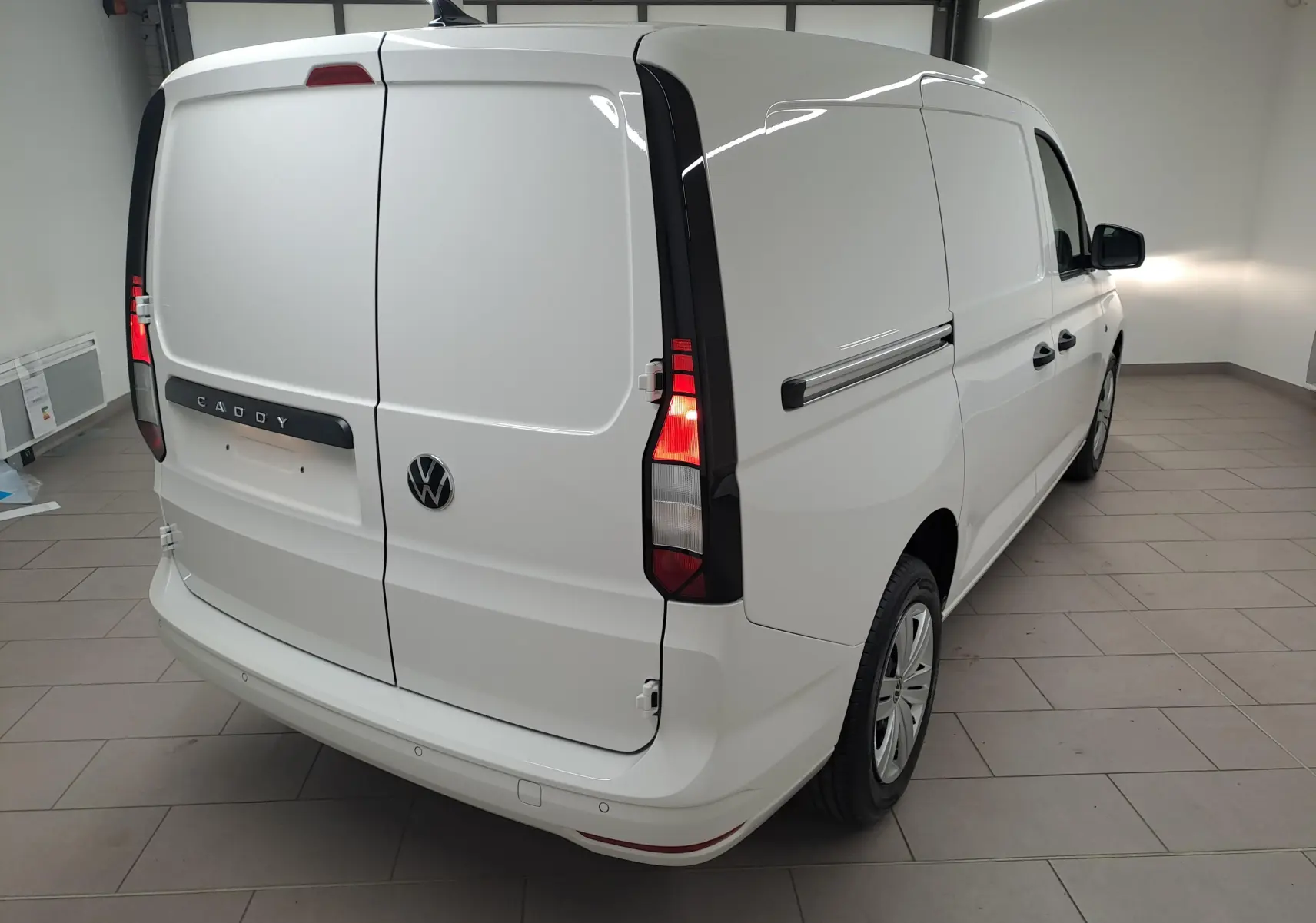 Vue 3/4 arrière droite du Volkswagen Caddy Cargo Maxi blanc Candy avec feux arrière allumés dans un intérieur.