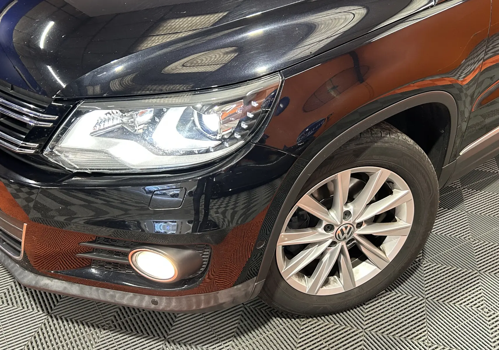 Vue 3/4 avant droit d’un Volkswagen Tiguan noir métal avec phare allumé et jante alliage visible.