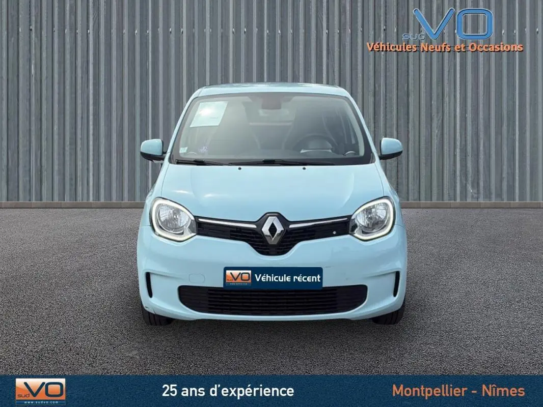 Renault Twingo III bleu clair vue de face, avec logo central et phares ronds allumés sur fond industriel.