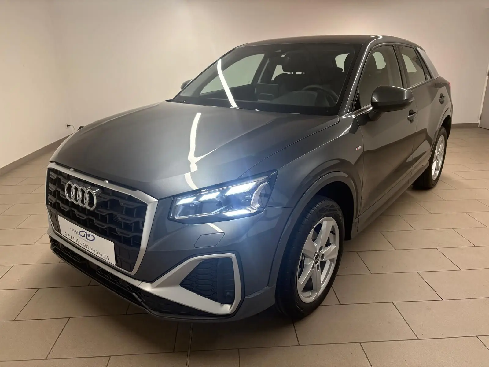 Audi Q2 35 TFSI S Line gris Daytona en 3/4 avant droit, phares allumés et calandre noire distinctive.