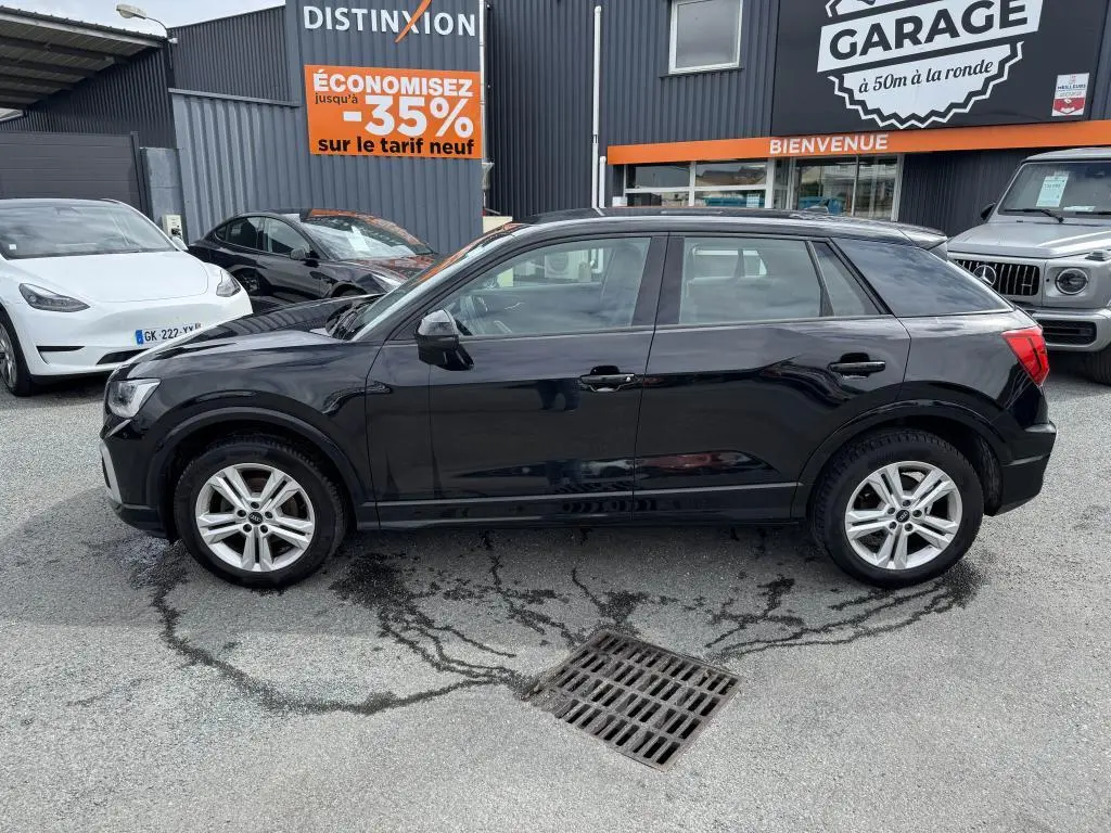Audi Q2 noire vue de profil côté gauche, garée devant un garage avec panneaux publicitaires visibles.