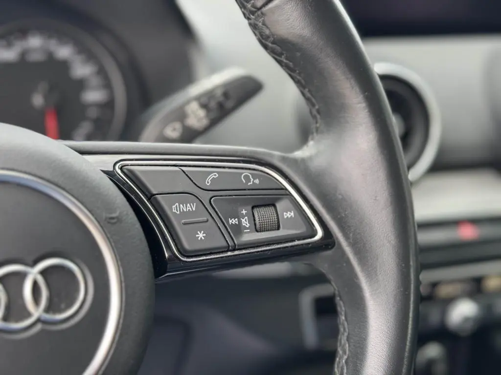 Gros plan sur les commandes gauche du volant cuir noir de l'Audi Q2 2022, avec tableau de bord flou en arrière-plan.