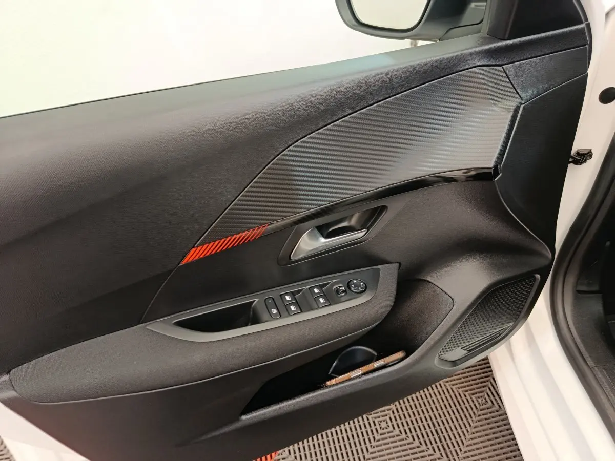 Vue intérieure côté gauche de la porte avant blanche d’une Peugeot 208 avec insert décor carbone et bande rouge.
