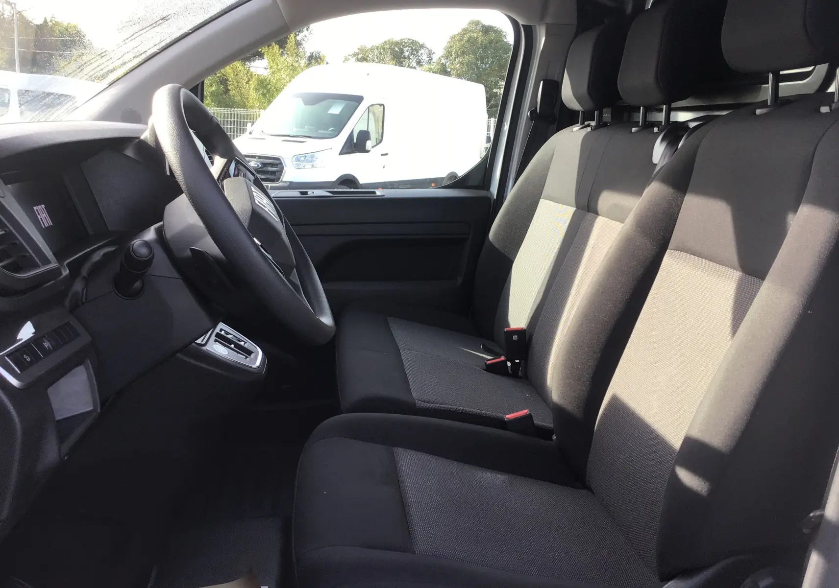 Vue intérieure côté conducteur du Fiat Scudo Fourgon blanc 2024, montrant les sièges avant en tissu noir et le volant.