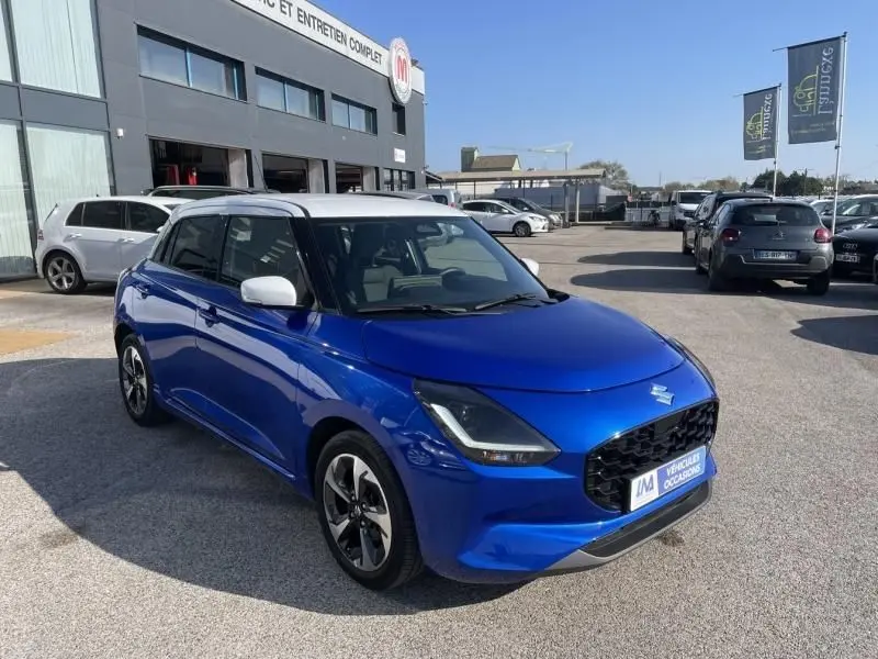 Suzuki Swift 2024 bleu métal vue 3/4 avant droit avec toit blanc et jantes bi-ton, stationnée en extérieur.