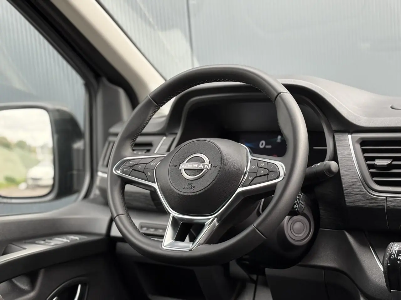 Volant Nissan en cuir avec commandes intégrées, tableau de bord noir et rétroviseur côté gauche visible.