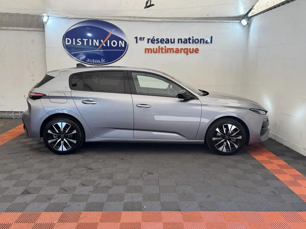 Profil droit d'une Peugeot 308 gris clair 2026 dans un showroom avec jantes noires et logo Distinxion en arrière-plan.
