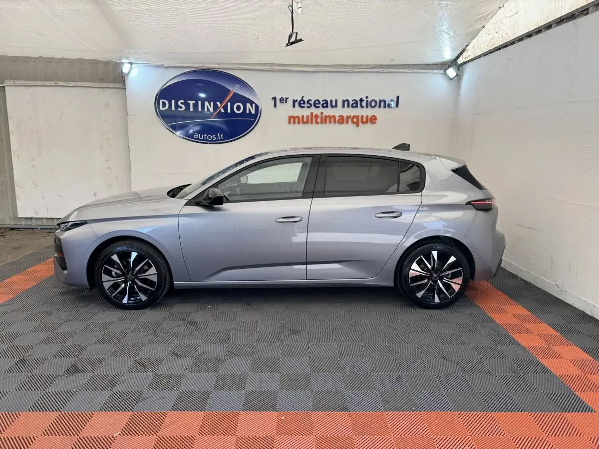 Vue de profil côté gauche d'une Peugeot 308 gris clair 2026 dans un showroom avec jantes noires et logo Distinxion en arrière-plan.