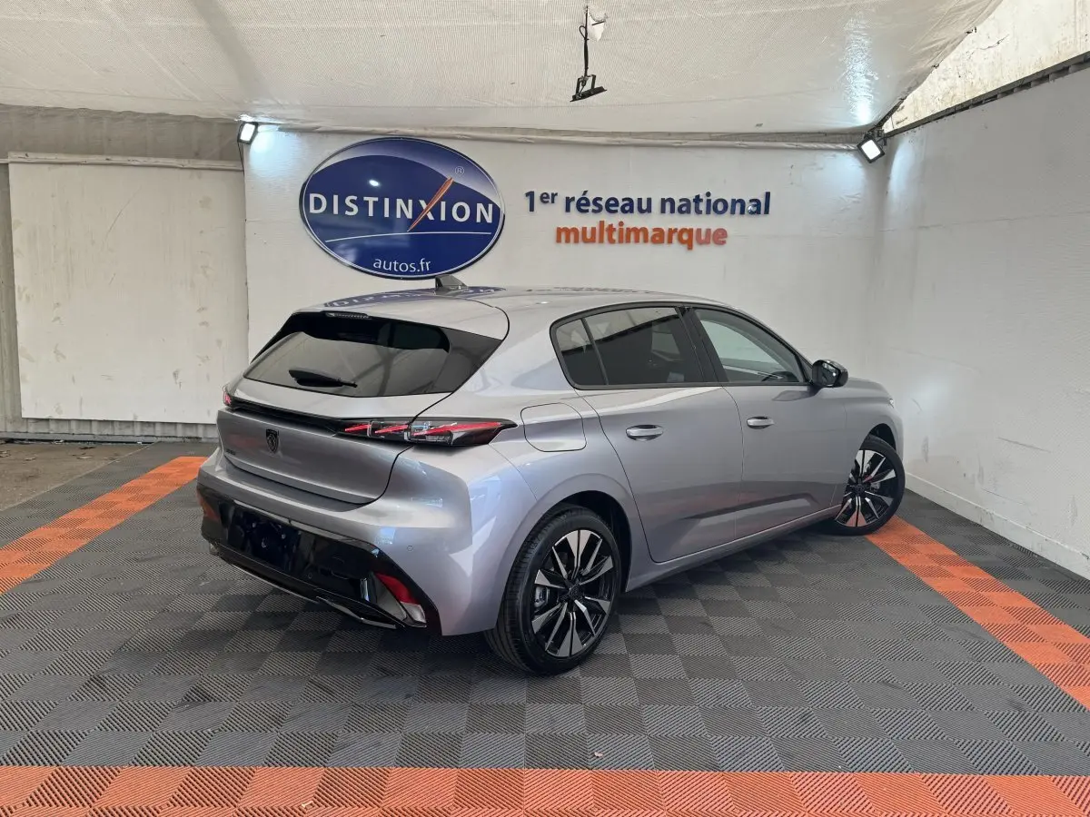 Vue 3/4 arrière droite d'une Peugeot 308 gris clair 2026 dans un showroom avec jantes noires et feux LED distinctifs.