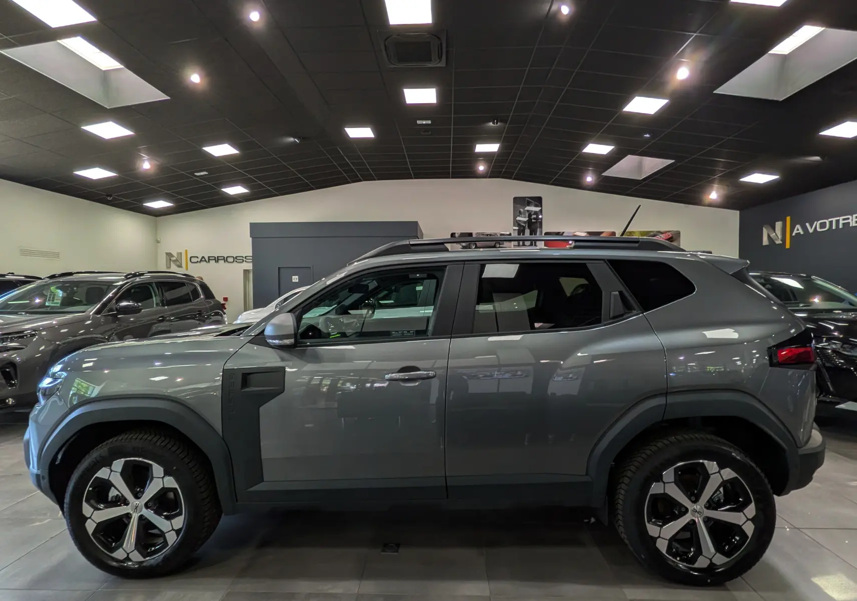 Vue de profil côté gauche du Dacia Duster Hybrid-G 150 CH 4x4 gris schiste avec jantes bi-ton et barres de toit en showroom.
