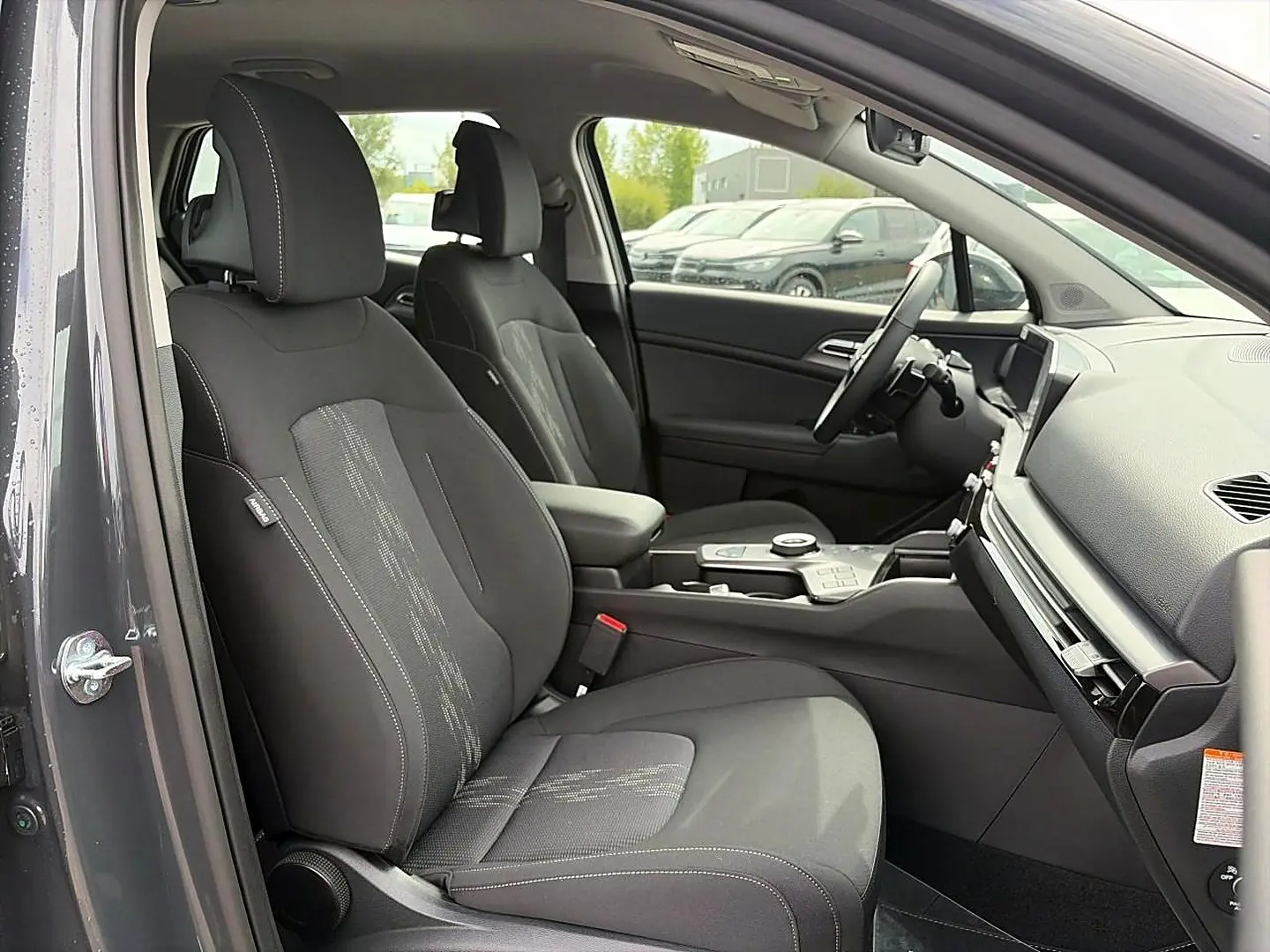 Vue intérieure côté conducteur du Kia Sportage 1.6 hybride gris eclipse, sièges tissu noirs avec surpiqûres blanches et console centrale moderne.