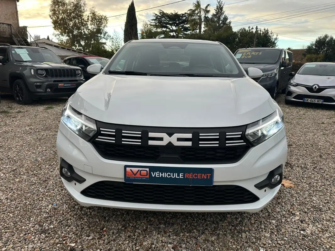 Vue frontale d'une Dacia Sandero blanche 2024 avec phares allumés et calandre noire distinctive.
