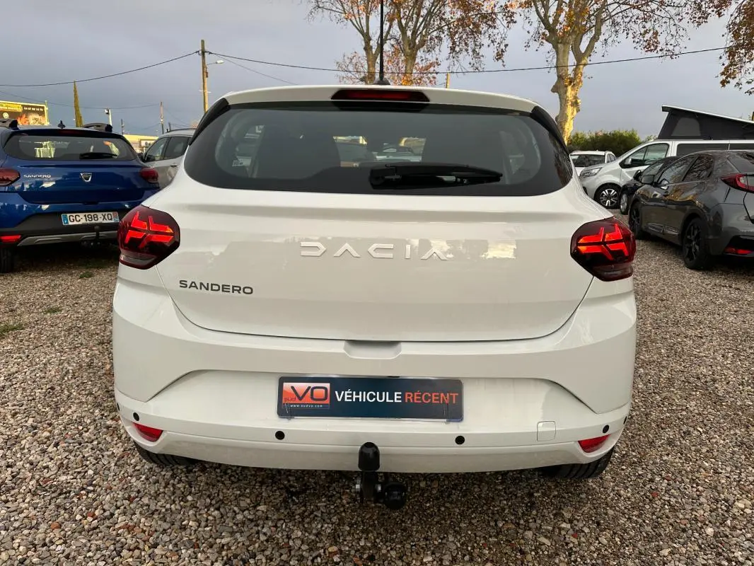 Vue arrière d'une Dacia Sandero blanche 2024 avec feux arrière allumés et attelage visible sur un parking.