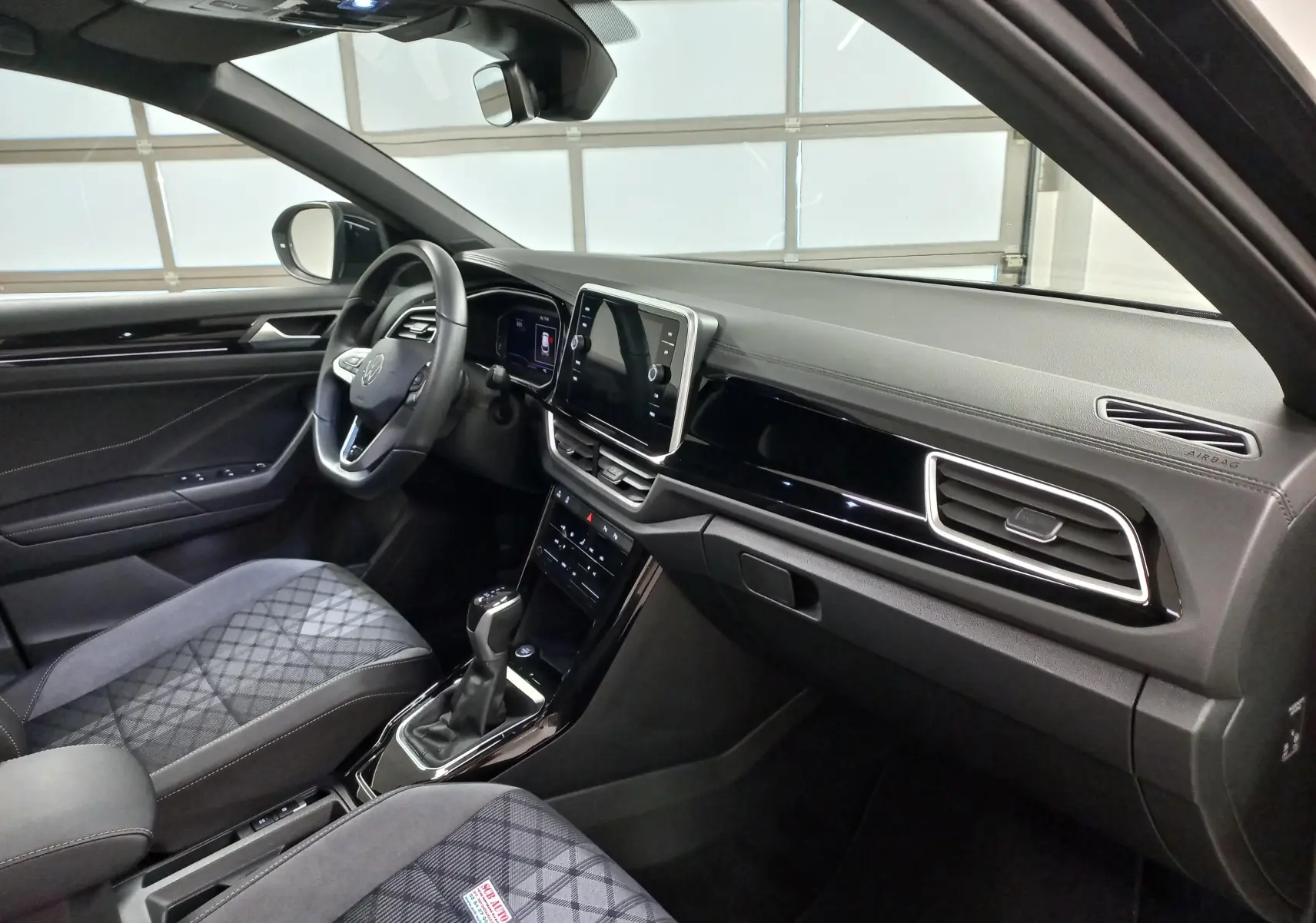 Vue intérieure côté conducteur du Volkswagen T-Roc 2025, tableau de bord noir avec écran tactile central et sièges tissu à motifs.